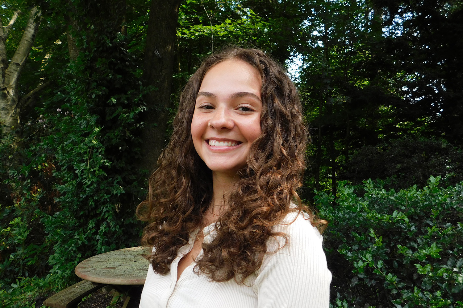 Josephine Burke, a young woman with long curly brown hair wearing an off-white long-sleeved shirt, smiles for photo in front of some trees and bushes.