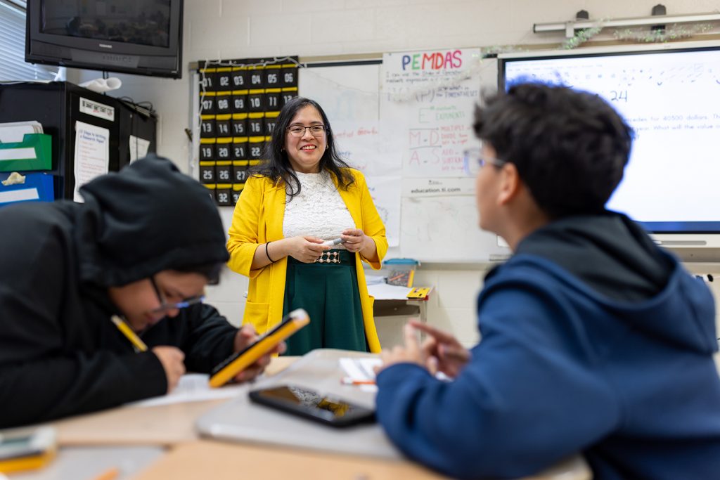 Female educator with yellow blazer speaks to two students in a classroom.