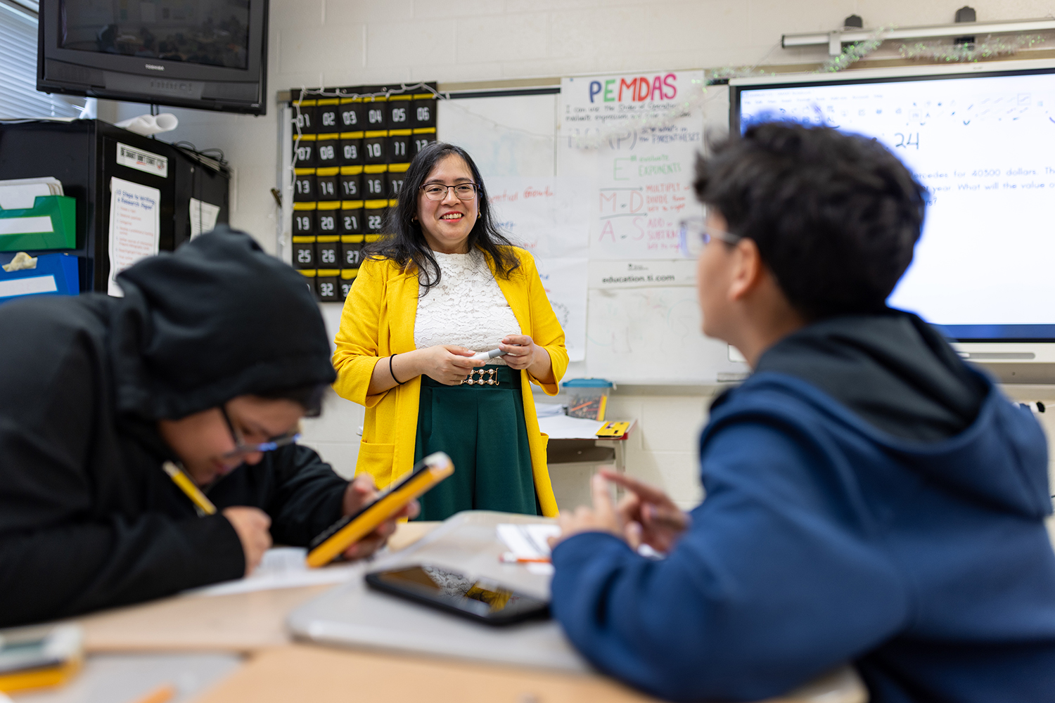 Female educator with yellow blazer speaks to two students in a classroom.