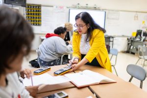 Female educator in yellow jacket shows a student a calculator in a classroom.