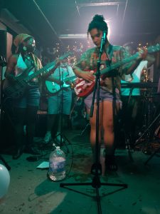 Members of a band holding instruments and singing into microphones.