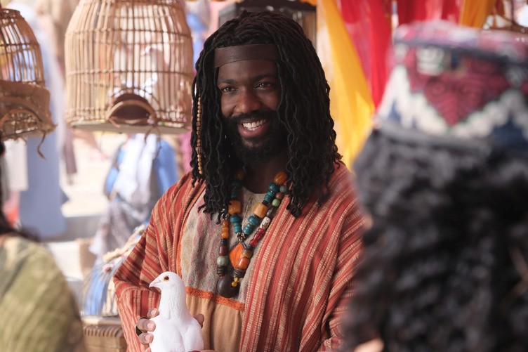 A man dressed in linen tunic with a bright colored necklace holds a white dove.
