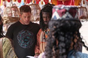 A man dressed in linen tunic with a bright colored necklace talks with a man in a black T-shirt.