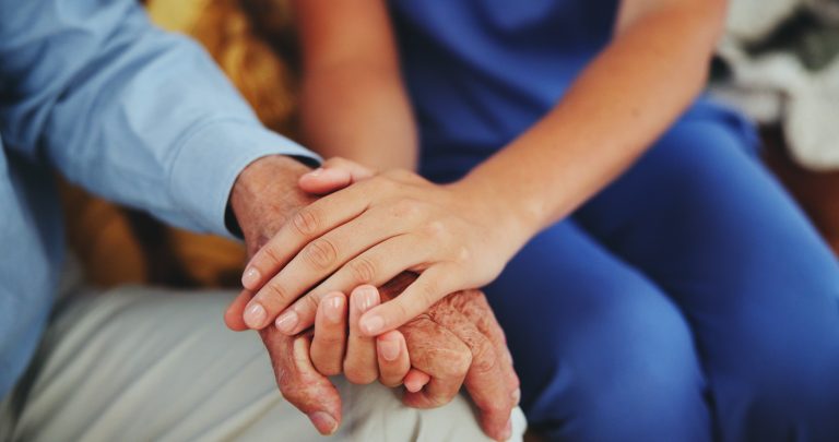 A younger person in hospital scrubs gently holds the hand of an older person