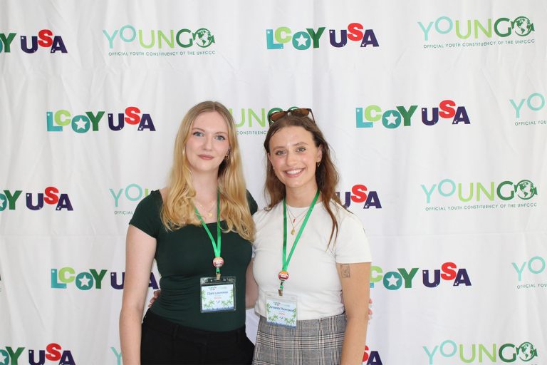 Two women posing for a photo in front of a backdrop that says LCOY USA