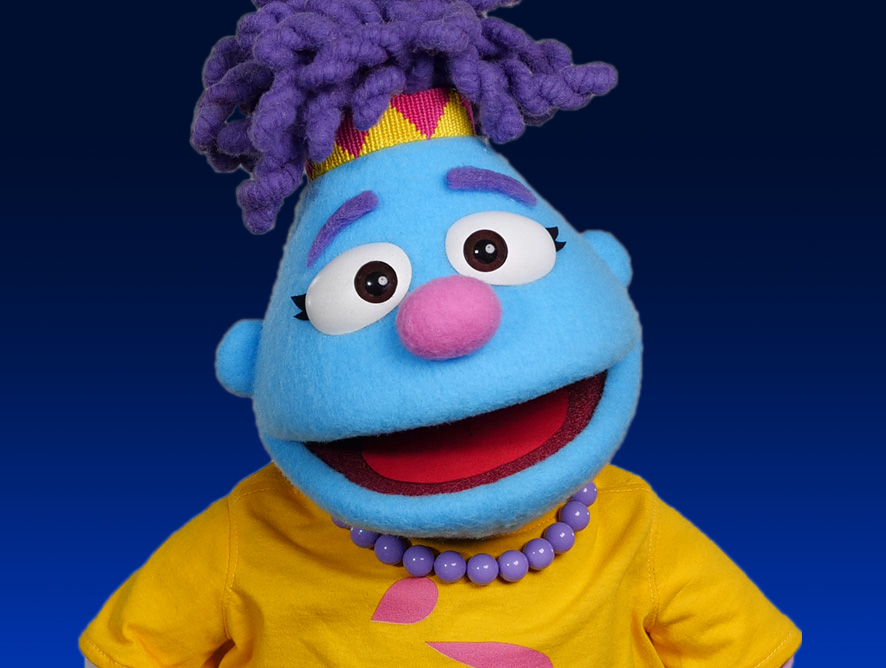 FYBS-hero-sm A blue puppet with a pink nose, white eyes with black pupils, and purple eyebrows is shown against a gradient blue background. The puppet has purple yarn hair styled in a high ponytail with a yellow and red checkered headband. It is wearing a yellow shirt and a necklace made of round purple beads.