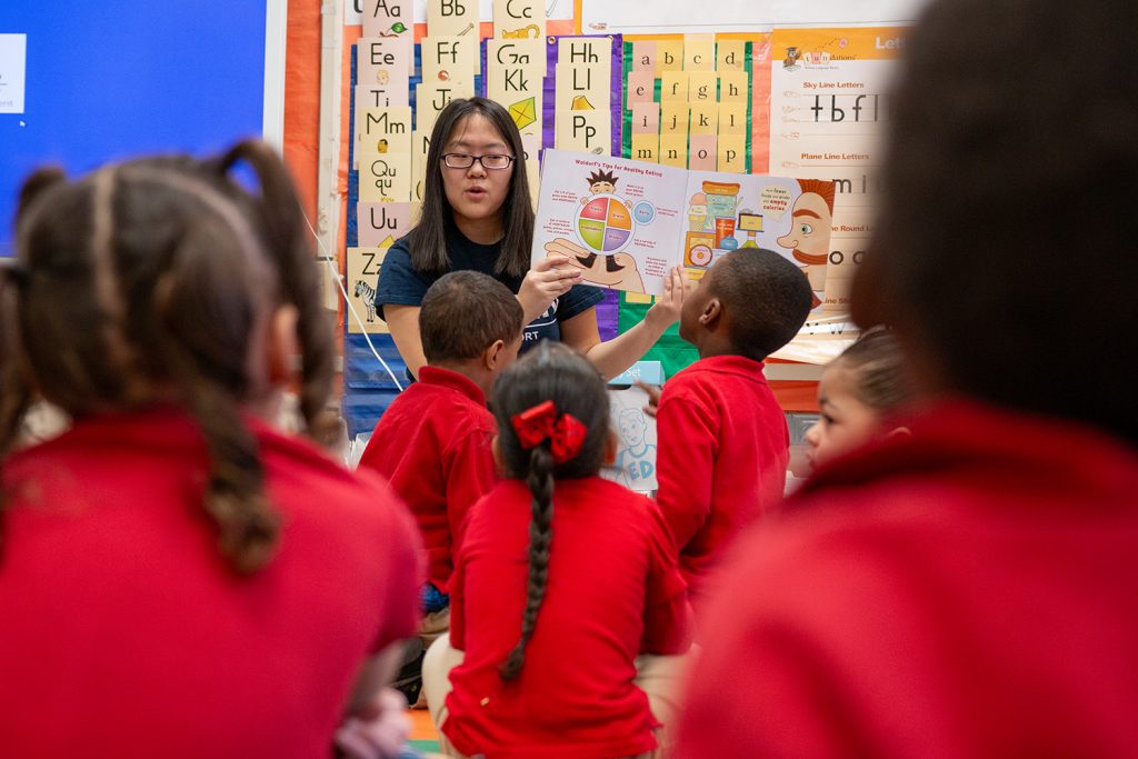 A female Asian American college student sits in front of a group of elementary school students, holding up a picture book and reading it.