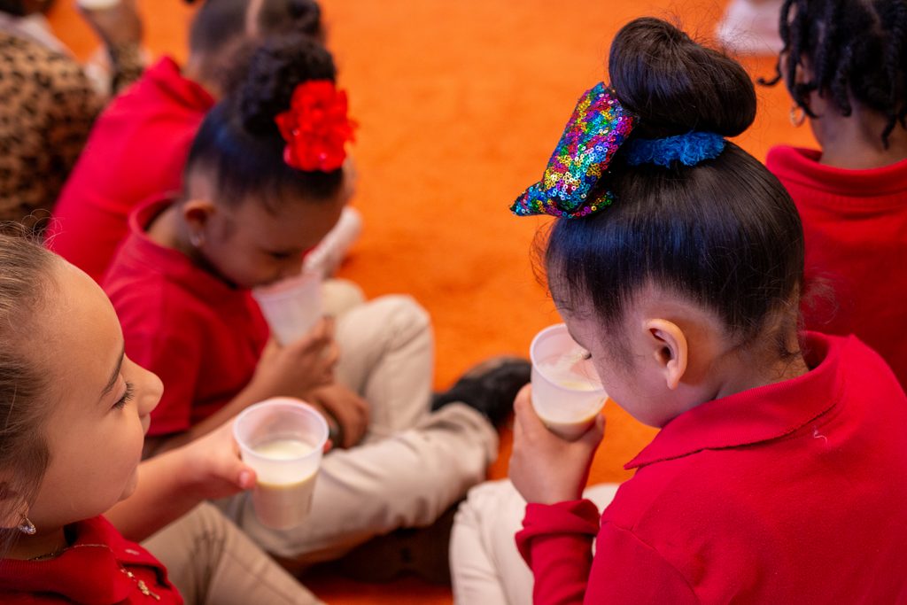 A group of elementary school students wearing red shirts sit on the floor while holding plastic cups filled with a smoothie. Two of the students smell the contents of their cups.