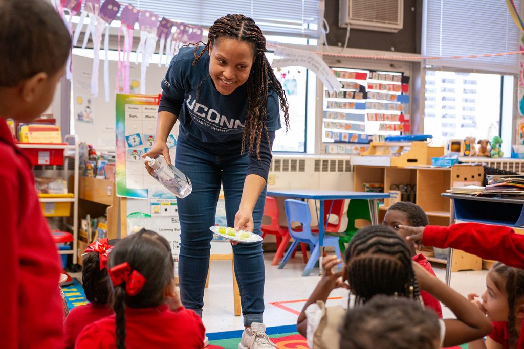 A Black female college student stands in an elementary school classroom and hands out pieces of food to students who are sitting on the floor.