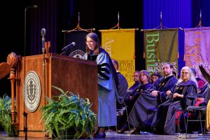 A woman with glasses standing at a podium in an academic robe with people in seats behind her also in academic robes