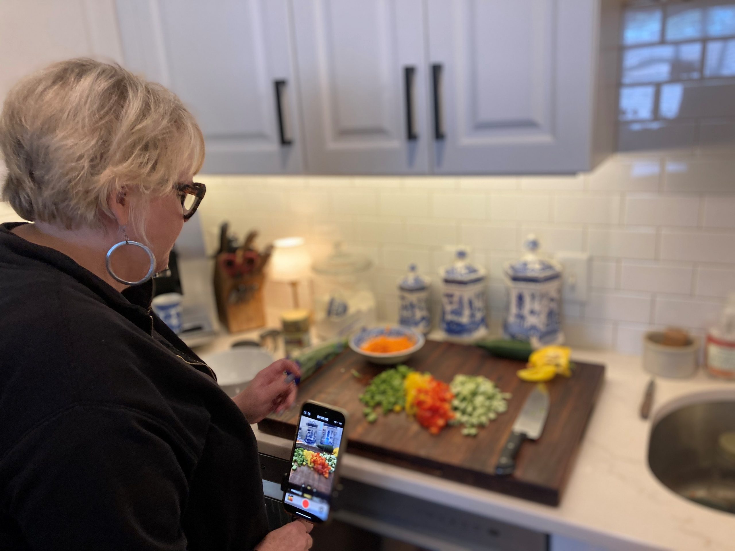 A woman stands in front of a kitchen counter with colorful cut vegetables on a cutting board.