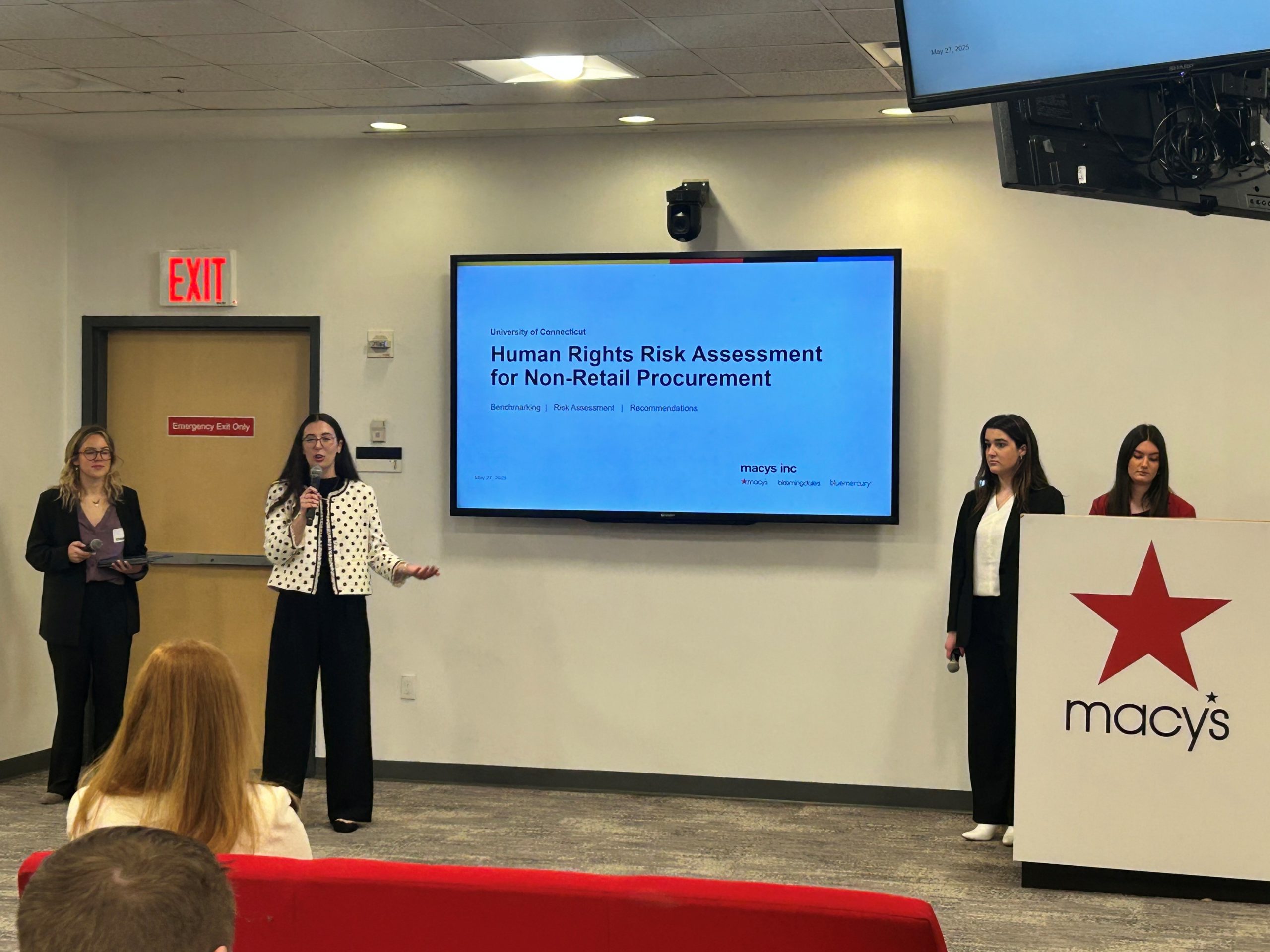 Four students give a presentation on a large screen, with a Macy's logo visible on a podium.