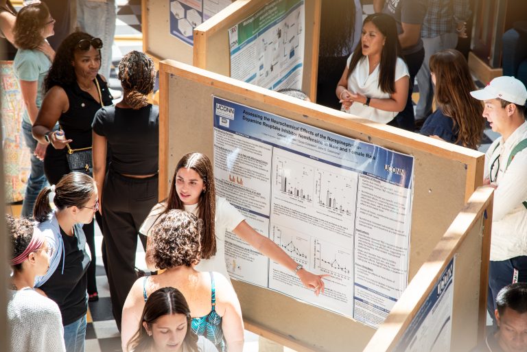 Students standing by posters on a large cork board talking to people about their research