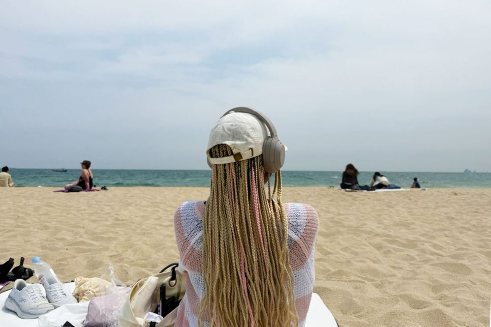 A woman with long blonde hair and a hat and headphones sitting on a blanket in the sand looking out to the ocean