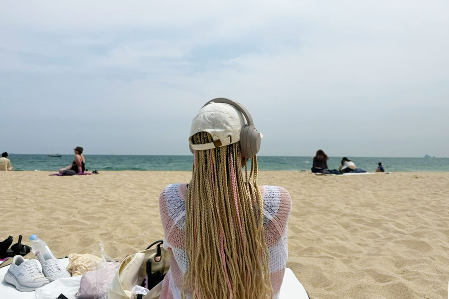 A woman with long blonde hair and a hat and headphones sitting on a blanket in the sand looking out to the ocean