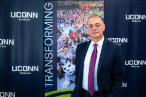 A man with glasses in a suit standing in front of a banner that says "UConn Research"