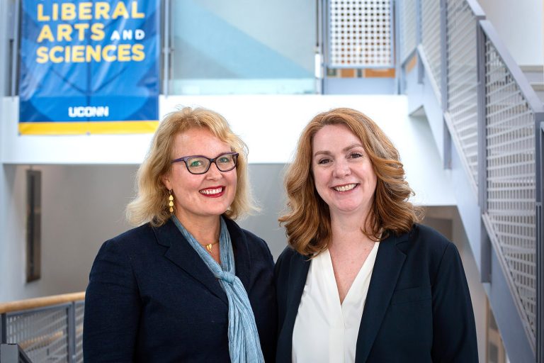 Manuela Wagner and Diane Quinn stand side-by-side in the entrance of the Austin Building with a Liberal Arts and Sciences banner in the background.