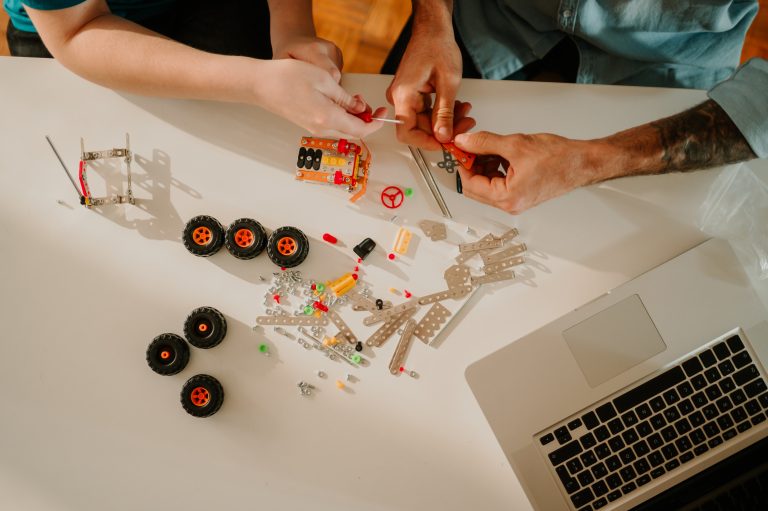 High angle shot of a white table with two people working with small toy car parts, and a laptop in the corner.