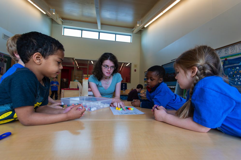 Student teacher works with elementary students at a school table.