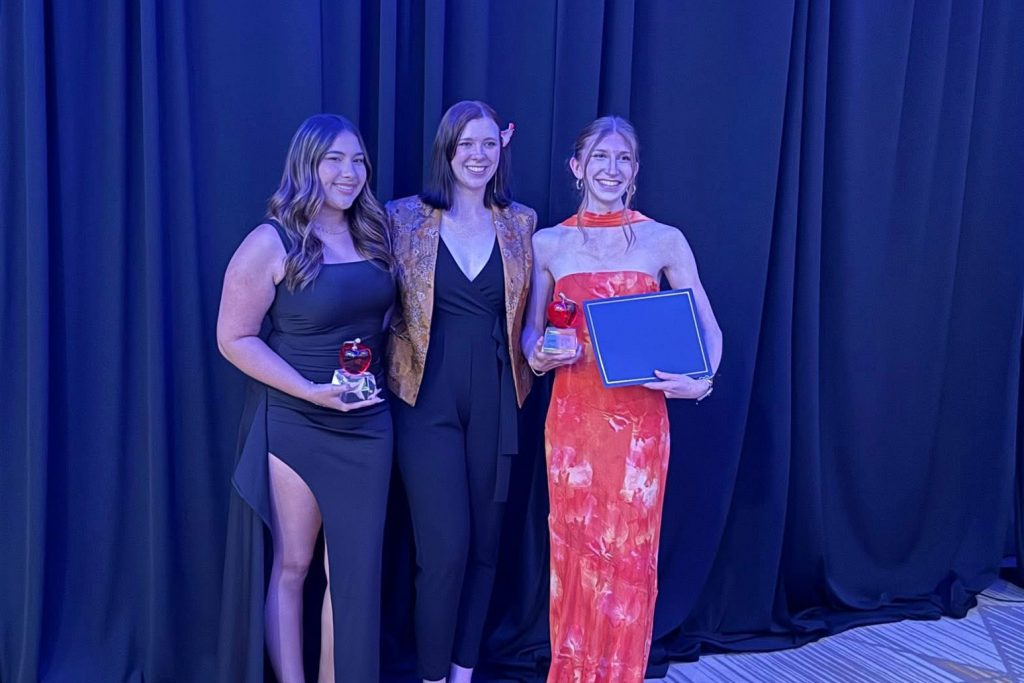Three women dressed in formal attire stand on stage with awards.