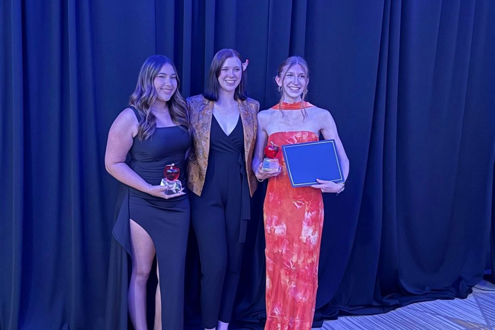 Three women dressed in formal attire stand on stage with awards.