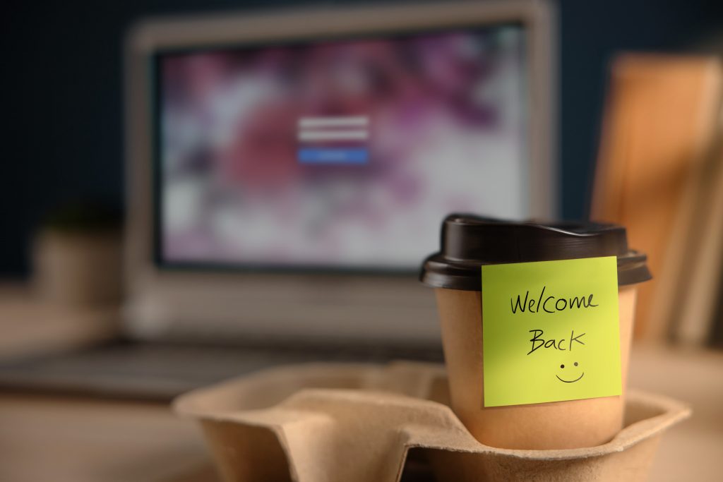 A takeout coffee cup on a desk with a post-it note that says "Welcome Back."