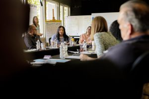 A group of people at a conference table listen to a woman talking.