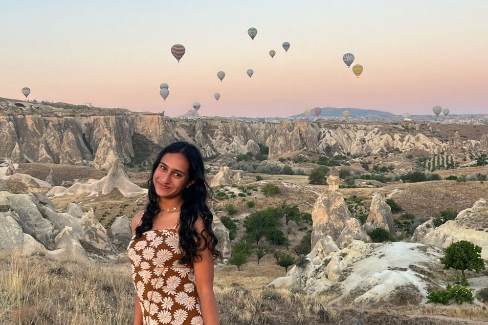 A woman with long dark hair standing with hot air balloons in the air behind her over a Turkish landscape