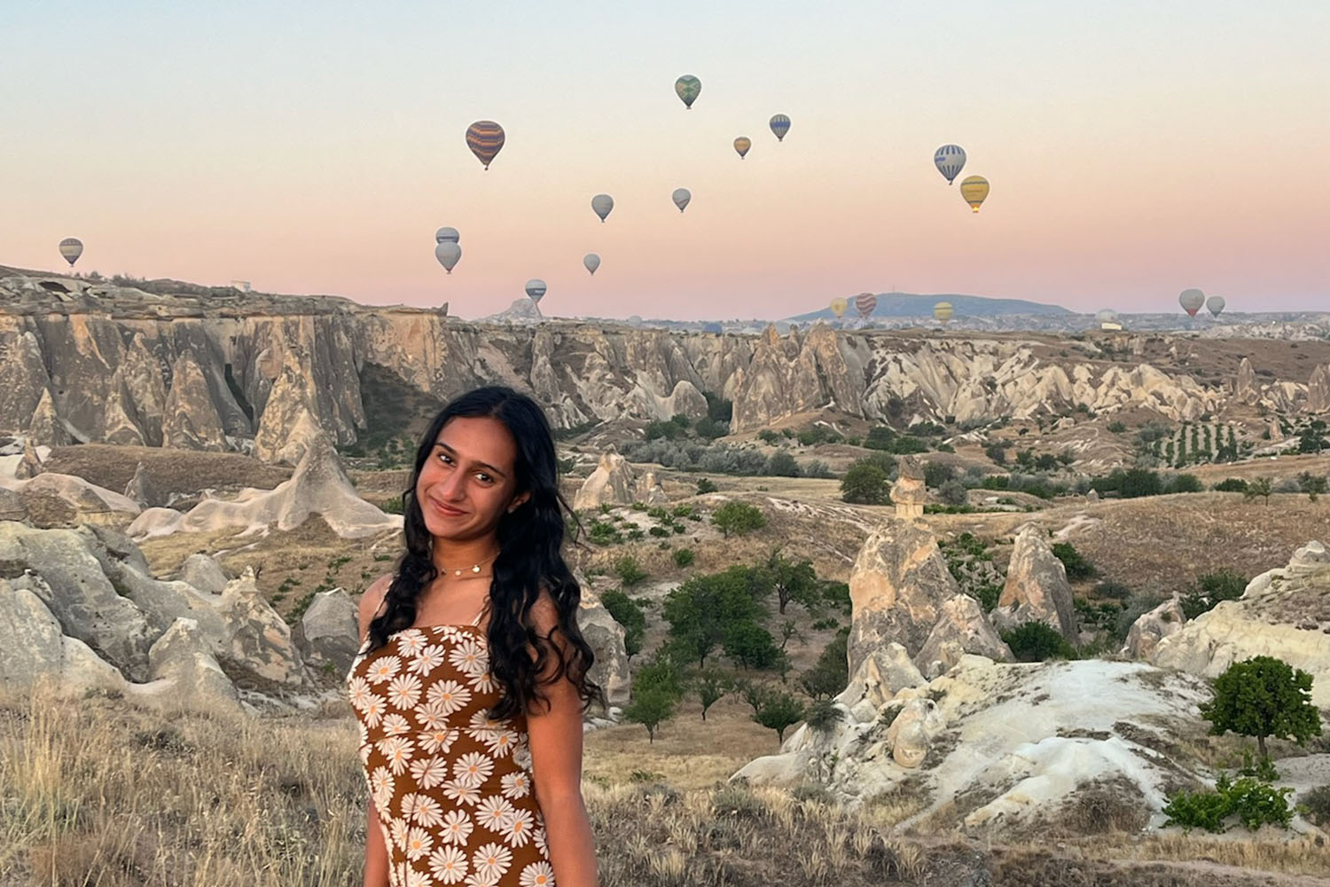 A woman with long dark hair standing with hot air balloons in the air behind her over a Turkish landscape