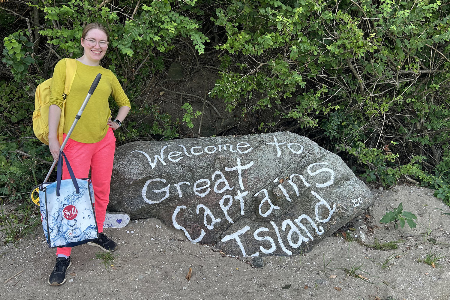 Hiis LaRose stands on a sandy beach next to a large rock that is half-submerged in the sand and has white words written on it that read "Welcome to Great Captains Island."