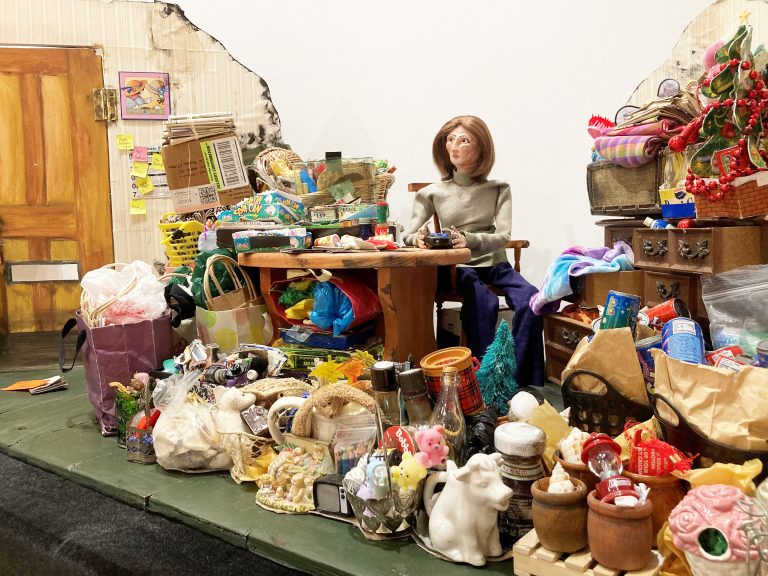 A puppet of a woman sitting at a table, surrounded by piles of strewn items.