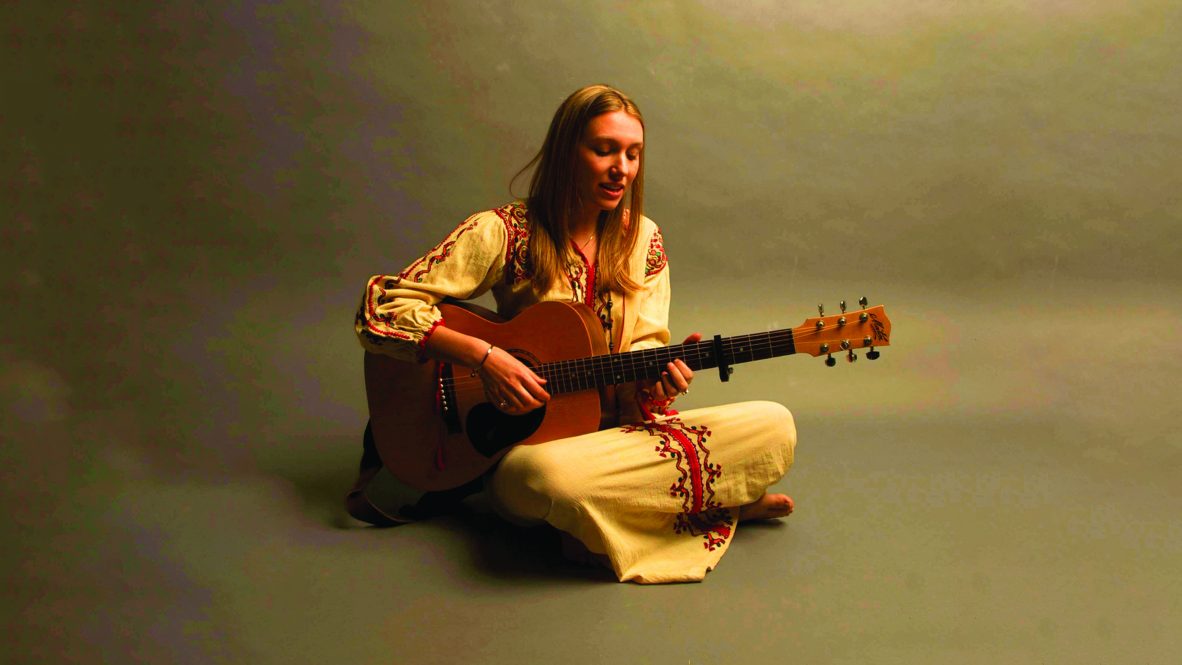 Actor and musician Rainee Blake sits on the ground holding an acoustic guitar.