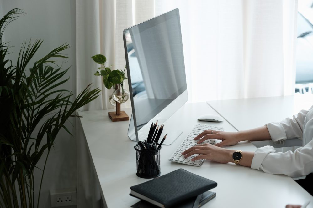 Person typing on a desktop computer at a clean, modern desk with plants and notebooks.