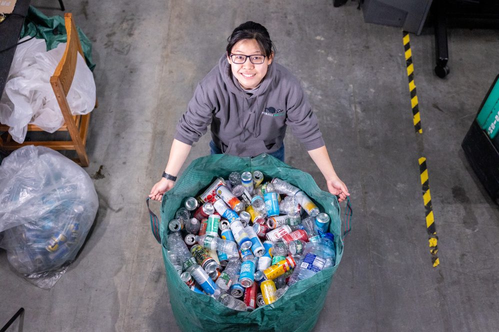 A student in a hoodie with a "Project Raccoon" logo poses with a large bin of recyclable plastic bottles