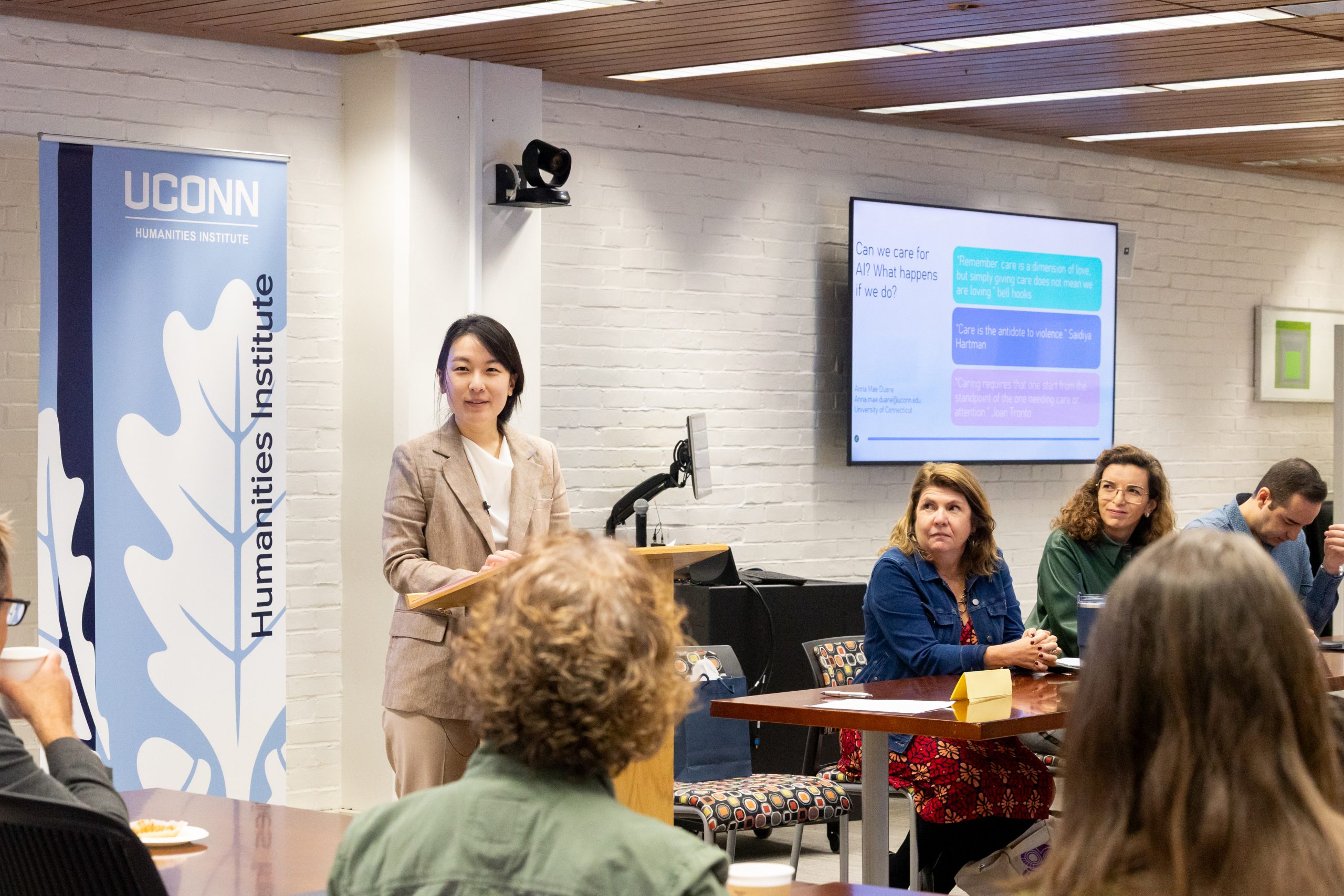 A professor addressing a room full of people with a banner displaying the UConn Humanities Institute logo visible behind her