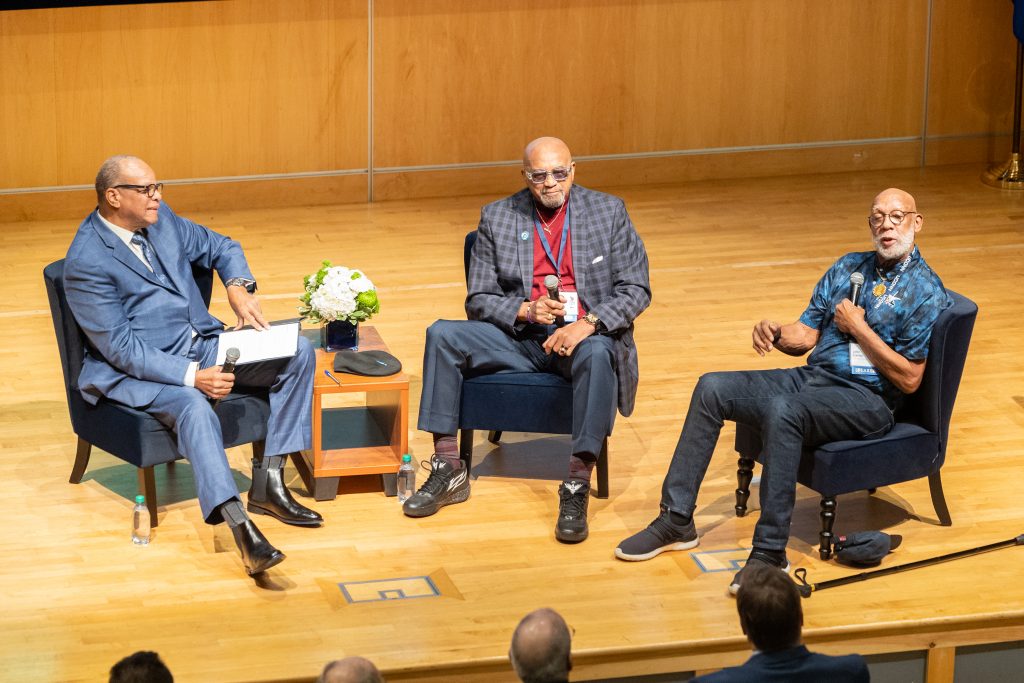 Three men sit on a stage during a discussion in front of an audience.
