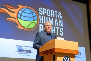 A man in a blue suit stands at a podium in front of a screen that says Sports and Human Rights.