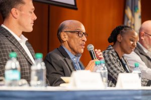 An older man with glasses speaks into a microphone while seated at a panel discussion.