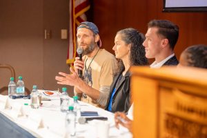 A man with a beard speaks into a microphone while seated at a panel discussion.