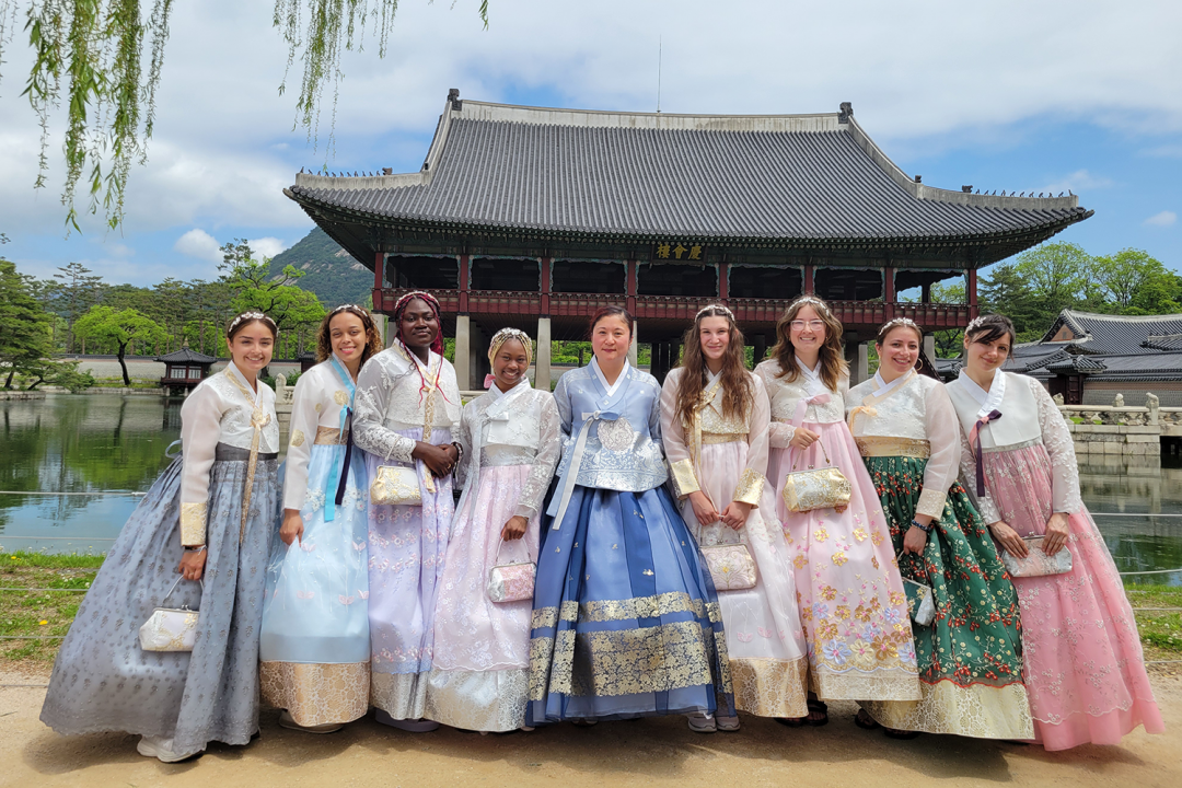 A diverse group of 9 women stands in front of a Korean palace wearing Hanbok, traditional Korean dress, in blues, purples, greens, and pinks.
