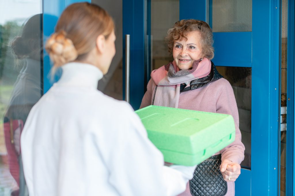 An older woman stands in front of a blue door receiving a meal delivery from a volunteer in a white jacket.