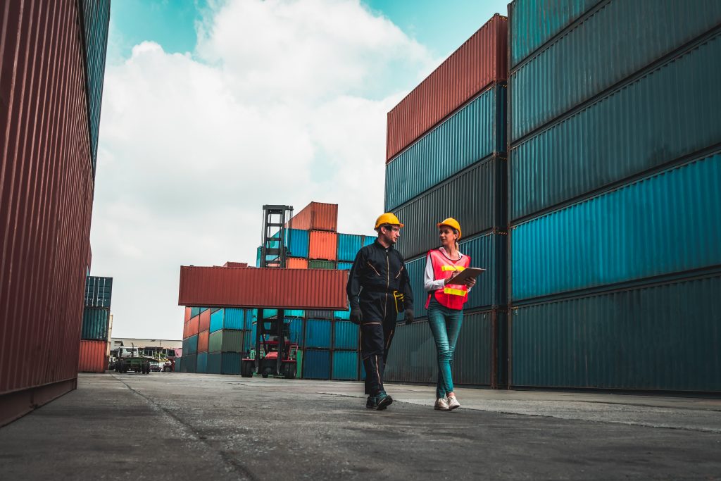 Two workers in hard hats walk side by side surrounded by shipping containers.