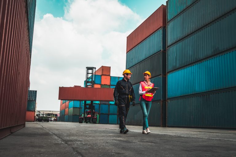 Two workers in hard hats walk side by side surrounded by shipping containers.