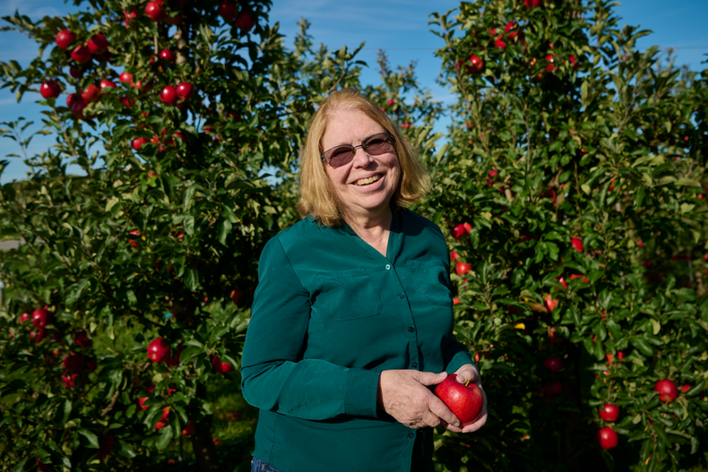 Susan Brown wears a green button-down top and smiles while holding a bright red apple in front of apple trees full of more bright-red apples on a blue-sky day.