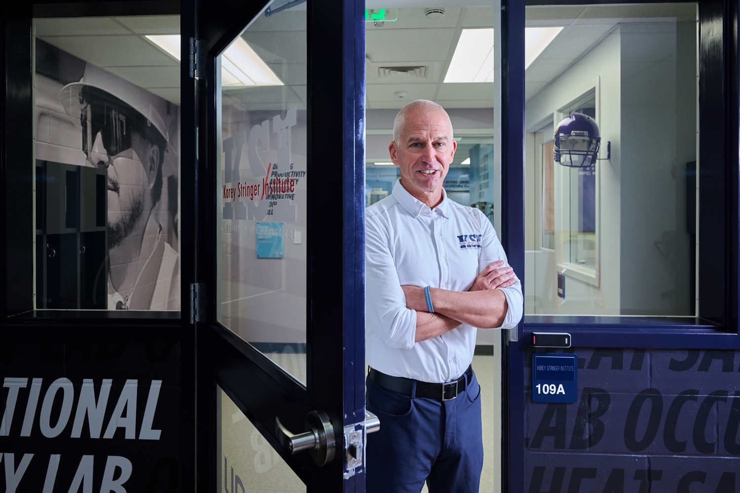 Person standing in the doorway of a lab labeled “109A,” wearing a white UConn-branded shirt and dark pants.