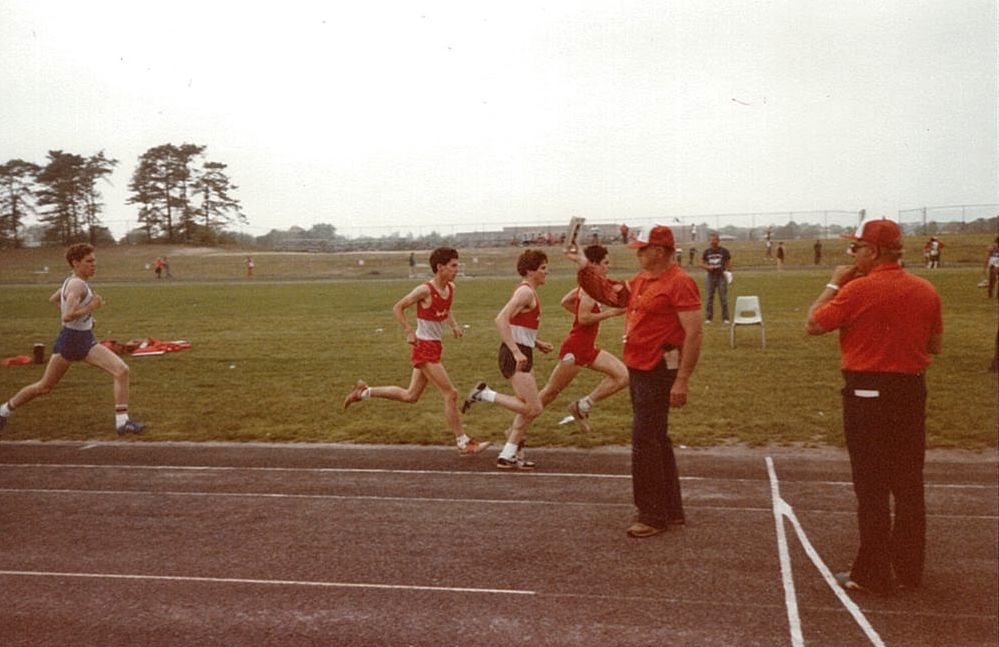 Four runners on a track during a race, three wearing red uniforms and one in blue. Two officials in red shirts and hats stand near the finish line.