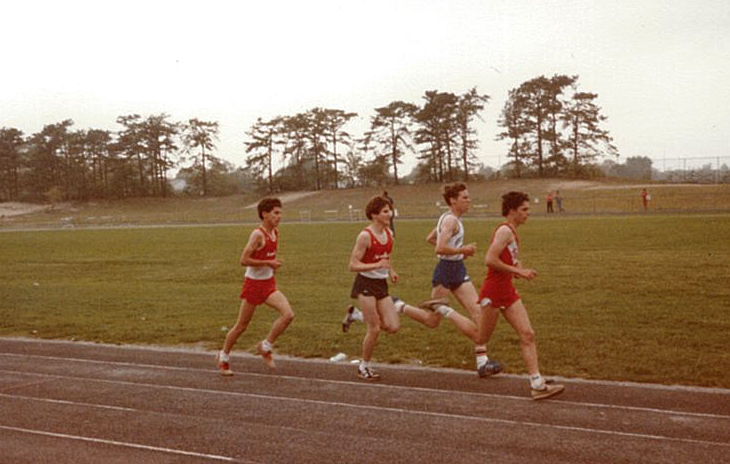 Four runners competing on a track during a race. Three runners wear red uniforms, and one wears a white top with blue shorts.