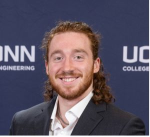 Headshot of a smiling man in a suit against a UConn Engineering backdrop.