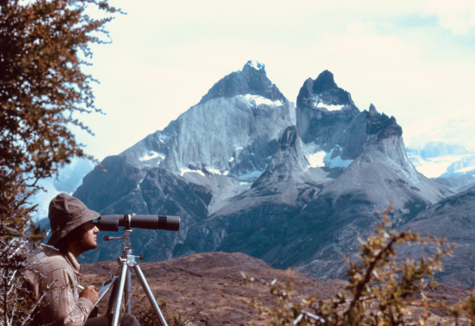 Morty Ortega, in a brown hat and plaid shirt at the left of the frame, looks through a telescope with a craggy mountain peak behind him at Torres del Paine National Park in Patagonia, Chile, in 1980