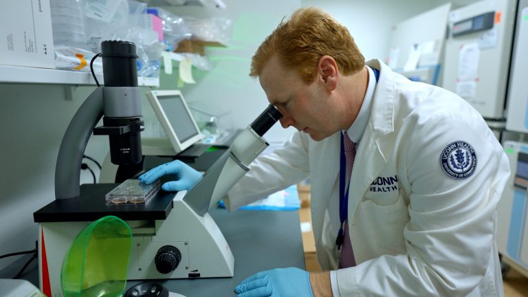 A man in a white lab coat looking through a microscope in a lab